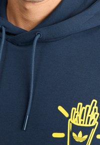 Navy sininen kangashuppari, jossa on etutasku, jossa on keltainen grafiikka ranskalaisista perunoista astiassa ja Adidas-logo-korostus.