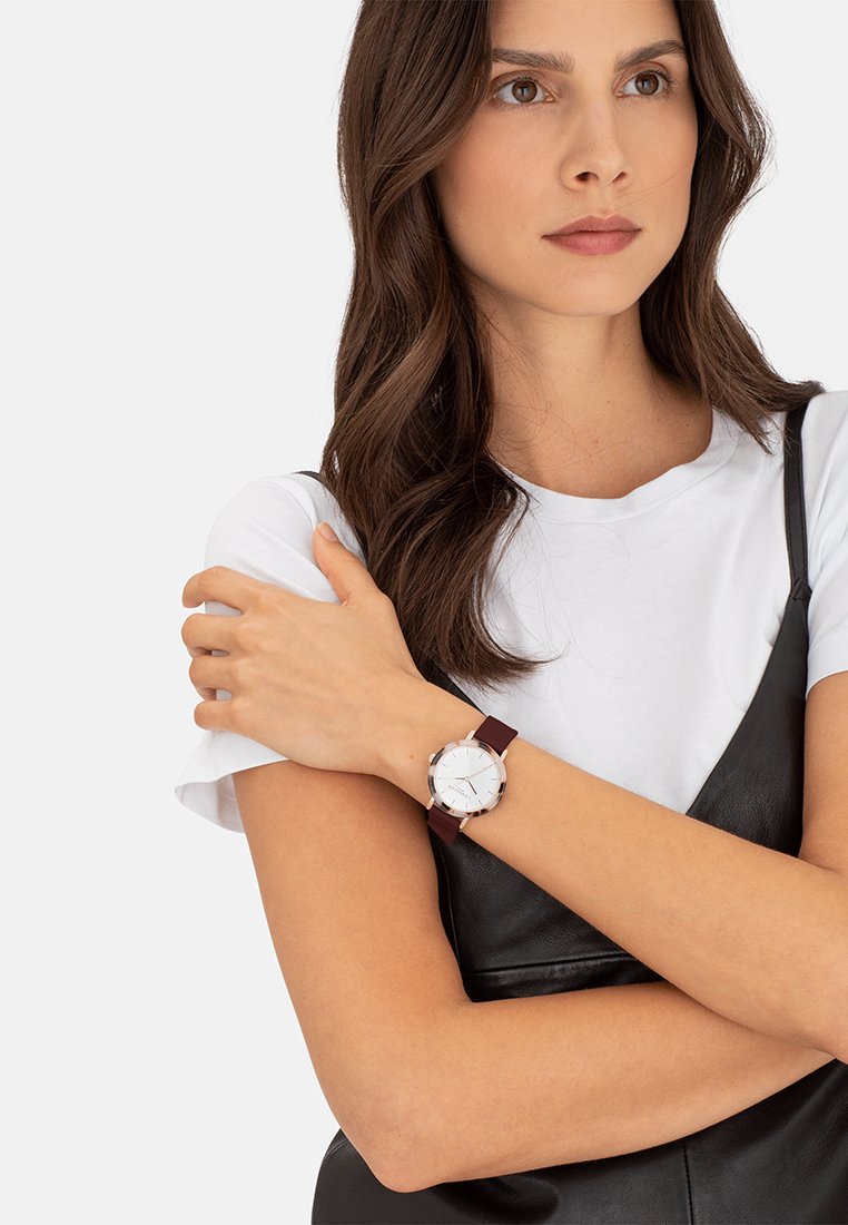 Runde Uhr mit silbernem Zifferblatt, weißen Stundenmarkierungen und burgunderfarbnem Lederarmband. Am Handgelenk getragen, kombiniert mit einem weißen Hemd und einem schwarzen Kleid.