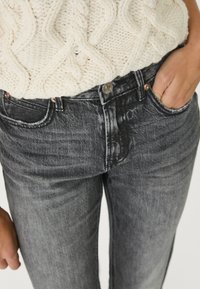 Jeans grigio scuro caratterizzati da un classico design a cinque tasche con chiusura a bottone e una leggera texture sbiadita. Indossati con un maglione crema texturizzato.