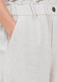 Pantaloni in lino beige chiaro con finitura texturizzata, cintura elastica, tasche laterali e dettagli con bottone marrone.