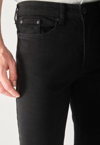 Mano masculina descansando cerca del bolsillo delantero de unos jeans de mezclilla negros ajustados contra un fondo claro.