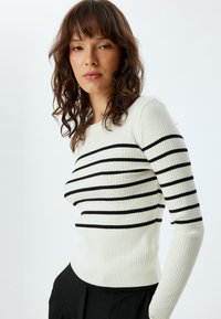 Maglione a costine di colore avorio con strisce orizzontali nere. Design aderente con maniche lunghe e scollo rotondo. Texture liscia.