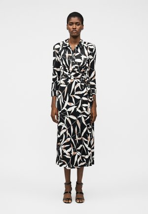 Woman standing wearing a black wrap dress with white and beige leaf patterns and black strappy heels on a plain light background.