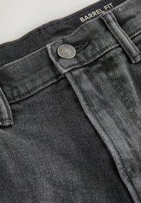 Gros plan sur la ceinture d'un jean en denim gris avec un bouton métallique portant l'inscription « Premium Denim Goods » et « Barrel Fit » imprimée à l'intérieur de la ceinture.