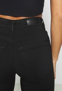 Jeans negros con una textura suave, corte ajustado y dos bolsillos traseros. Presentan un detalle de costura y una etiqueta de cuero con la marca en la cintura.