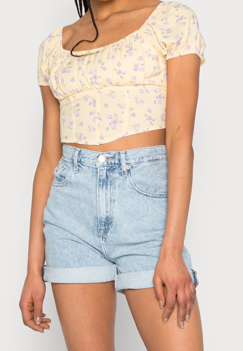 Haut court à fleurs jaune avec manches bouffantes, assorti à un short en jean bleu clair taille haute, avec revers roulés et fermeture à boutons sur le devant.