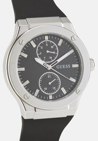 Reloj en tono plateado con una correa de goma negra y un dial negro. Las características incluyen sub-diales de hora, minuto, día y fecha, marcadores de horas y manecillas luminiscentes.
