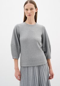 Grauer Pullover mit rundem Halsausschnitt und Dreiviertelärmeln, mit einer glatten Textur. Kombiniert mit einem grauen Faltenrock.