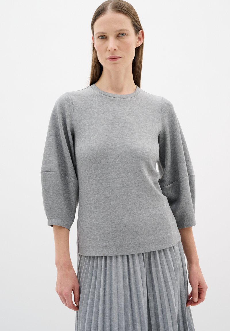 Grauer Pullover mit rundem Halsausschnitt und Dreiviertelärmeln, mit einer glatten Textur. Kombiniert mit einem grauen Faltenrock.