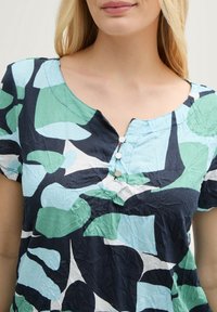Haut à manches courtes en tissu froissé présentant un motif abstrait bleu, vert et blanc avec un col rond et trois boutons décoratifs.