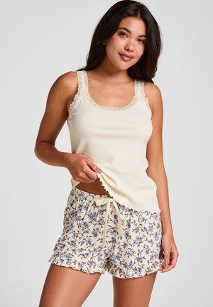 Femme portant un débardeur crème bordé de dentelle et un short à cordon floral, debout, tenant l'ourlet du débardeur d'une main.