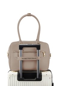 Borsa beige Victoria Hyde London fissata al manico di una valigia bianca con manico telescopico esteso.