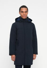 Mann in einer dunkelblauen Winterparka mit Kapuze, mit Reißverschluss und Klappenverschluss, steht vor einem schlichten weißen Hintergrund.