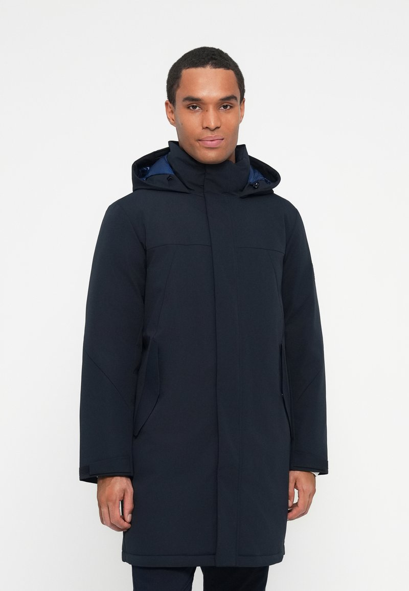 Mann in einer dunkelblauen Winterparka mit Kapuze, mit Reißverschluss und Klappenverschluss, steht vor einem schlichten weißen Hintergrund.