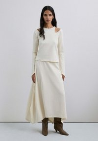 Ensemble crème comprenant un haut à manches longues avec découpes aux épaules et une jupe maxi fluide et asymétrique, associée à des bottes à talons marron.