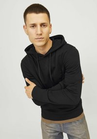 Black hoodie made of soft fabric with a drawstring hood, ribbed cuffs, and a kangaroo pocket, layered over a beige shirt and paired with jeans.