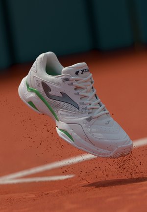 Zapatilla deportiva blanca con acentos grises y verdes, diseño de cordones, parte superior de malla texturizada y suela con patrón, suspendida sobre una pista de arcilla.