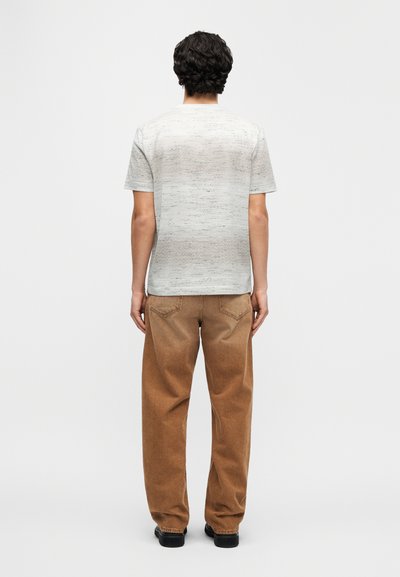 Ανοιχτό γκρι μπλουζάκι με διακριτική υφή και κοντά μανίκια, συνδυασμένο με καφέ παντελόνι από κοτλέ σε άνετη εφαρμογή. Ορατά μαύρα επίπεδα παπούτσια.