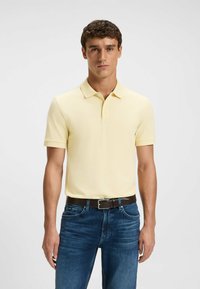 Man som står med en ljusgul kortärmad polo tröja nedstoppad i mörkblå jeans med ett brunt bälte mot en enfärgad bakgrund.