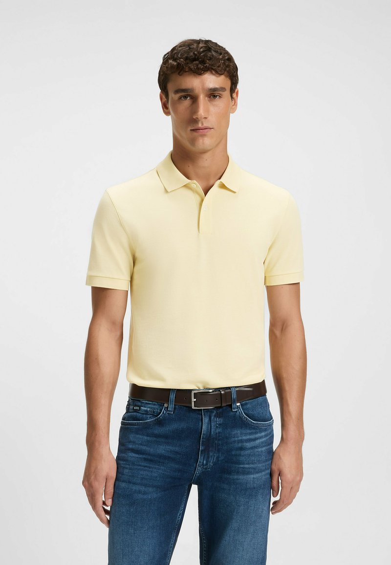 Man som står med en ljusgul kortärmad polo tröja nedstoppad i mörkblå jeans med ett brunt bälte mot en enfärgad bakgrund.