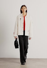 Hvid denimjakke med broderede detaljer, rød skjorte under, stribede sorte bukser, sort taske og sorte sko, model der står foran en neutral baggrund.