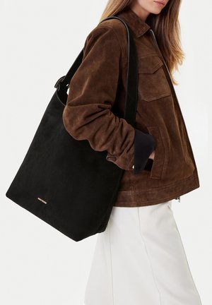 Mujer con chaqueta de ante marrón y pantalones blancos, lleva un gran bolso tote negro con asas al hombro en el brazo.