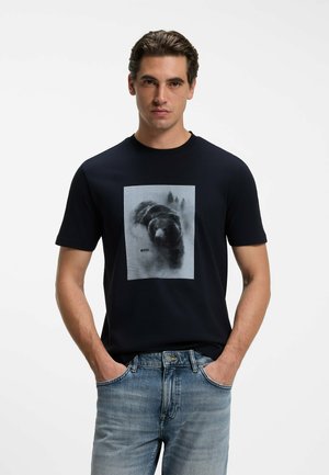 Man wearing a black t-shirt with a black and white bear graphic and light blue jeans, standing with hands in pockets against a white background.