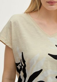 Beige V-hals top met korte mouwen, met een zwart en grijs bloemenpatroon. Zachte textuur met een lichte, slubbed stofafwerking.