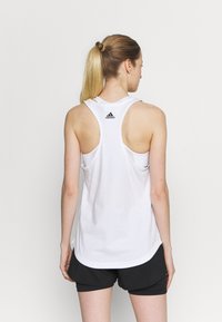 Canotta atletica bianca con scollatura rotonda e design a schiena scoperta. Presenta un piccolo logo Adidas nero sulla parte superiore della schiena e una vestibilità ampia.