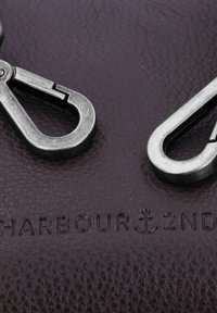 Superficie de cuero oscuro con dos clips metálicos, con un patrón texturizado. 'HARBOUR 2ND' grabado abajo, símbolo de ancla visible como acento.