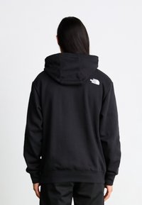 Personne aux longs cheveux foncés portant un sweat à capuche noir avec un logo blanc sur la partie supérieure droite du dos.