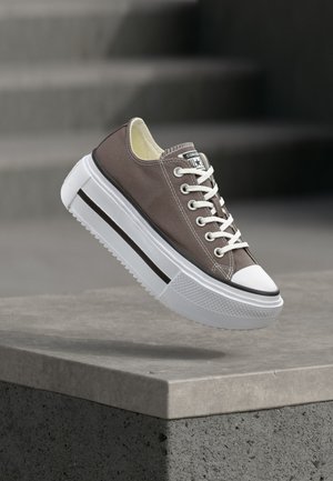 Brązowy niski sneaker z białymi sznurówkami i grubą białą podeszwą, unoszący się nad betonową krawędzią na zewnątrz.