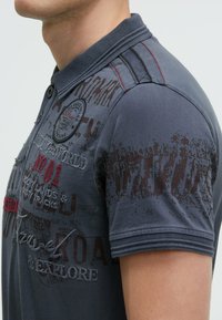 Dark gray polo shirt featuring front text graphics in red and brown, textured collar, and narrow stripes on the shoulders.
