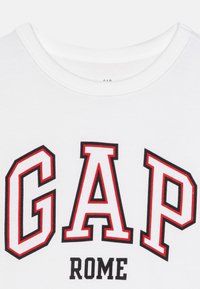 Tricou alb cu decolteu circular, având inscripții îndrăznețe în negru și roșu "GAP" și "ROME" într-un font sportiv. Fabric din bumbac moale.