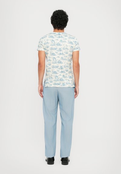 Lichtblauw met patroon t-shirt op een crème achtergrond, gecombineerd met lichtblauwe streepbroek. Model is van achteren te zien en draagt zwarte schoenen.