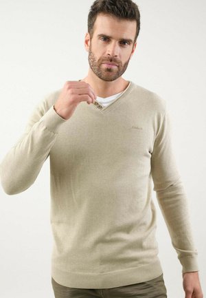 Pull-over beige en V en tissu doux, arborant un logo discret sur la poitrine et des poignets et un ourlet côtelés. Détail de boutons visible au niveau de l'encolure.