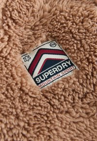 Syntetyczne futro w jasnobrązowym kolorze z teksturowaną powierzchnią. Posiada prostokątną łatkę z tkaniny z logo "Superdry Mountain Sport".