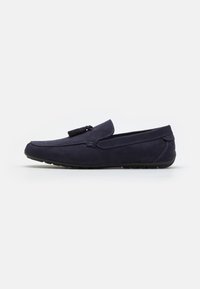 Mocassin en daim bleu marine avec détail de glands, style à enfiler, et semelle en caoutchouc noire, présenté en profil latéral sur fond blanc.