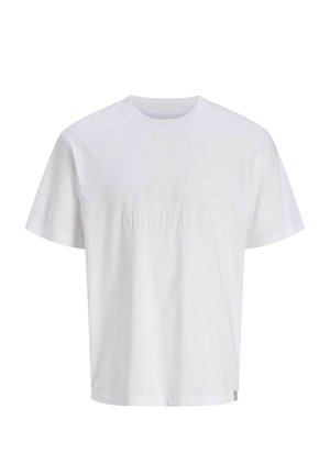 Smilende dreng iført en sort-hvid ternet skjorte over en hvid T-shirt, løsthængende blå jeans og beige sko, der står på en ensartet baggrund.