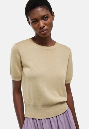 Femme portant un haut en tricot beige à manches courtes et une jupe lavande, debout devant un fond gris clair uni.