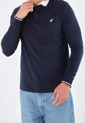 Homme portant un polo à manches longues bleu marine avec col et poignets blancs, jean bleu clair, main ajustant le col.