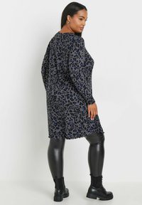 Langärmliges Kleid aus einem grauen Stoff mit einem Blau- und Schwarz-Leopardenmuster. Abgerundeter Saum, lockere Passform, kombiniert mit schwarzen Leggings und Stiefeln.