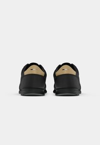 Paire de chaussures de sport noires avec des languettes beiges au talon, vues de dos sur un fond gris clair uni.