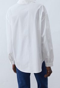 Camicia bianca a maniche lunghe con colletto, caratterizzata da una vestibilità comoda e maniche con polsini, abbinata a jeans blu scuro. Tessuto dalla texture liscia.
