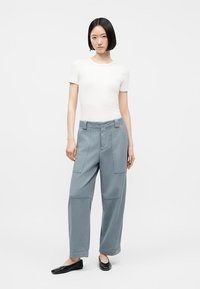 Γυναίκα που στέκεται μπροστά σε λευκό φόντο φορώντας λευκό τοπ με κοντά μανίκια, ανοιχτό μπλε παντελόνι με φαρδιά μπατζάκια και μεγάλες μπροστινές τσέπες, και μαύρα φλατ παπούτσια.