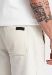 Pantalones de chándal de algodón beige con un bolsillo trasero que tiene una etiqueta "ombre" negra. Textura suave con detalles de costura visibles.