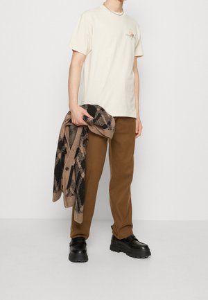 Mężczyzna w kremowej koszulce z krótkim rękawem, brązowych spodniach i czarnych butach, trzymający brązowo-czarny sweter w wzory na neutralnym tle.