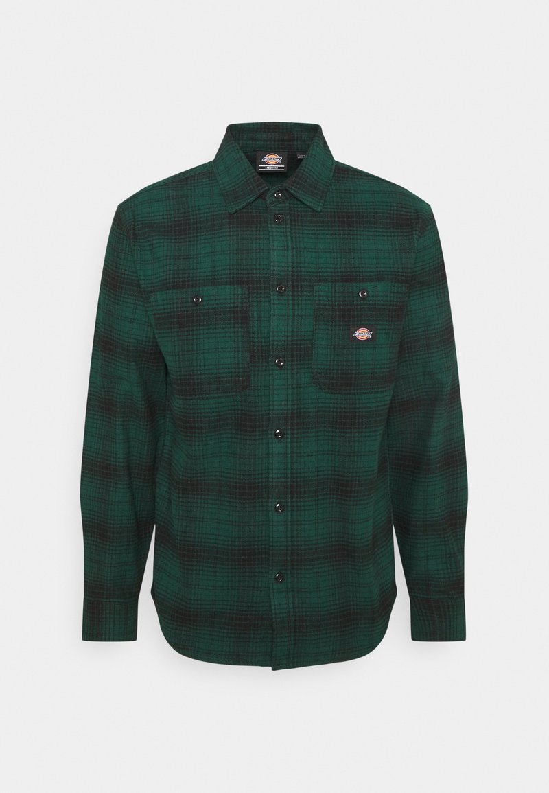 Chemise à manches longues à carreaux verts et noirs, boutonnée, avec deux poches poitrine à l'avant, dont une ornée d'un petit écusson circulaire.