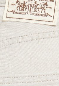 Bela tkaninska oznaka z rjavim besedilom in grafiko, ki prikazuje dva konja in jahače. Oznaka vključuje "Patented May 20187" in blagovno znamko.