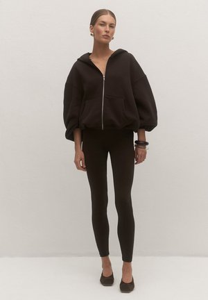 Kobieta ubrana w oversize czarną bluzę z kapturem na suwak, czarne legginsy i czarne szpilki, stojąca na jasnym, jednolitym tle.